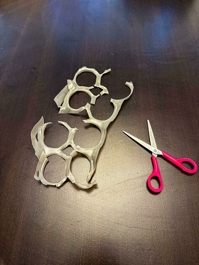 Plastic six-pack rings cut apart next to red-handled scissors on a dark wooden table, evoking nostalgic 90s DIY crafts.