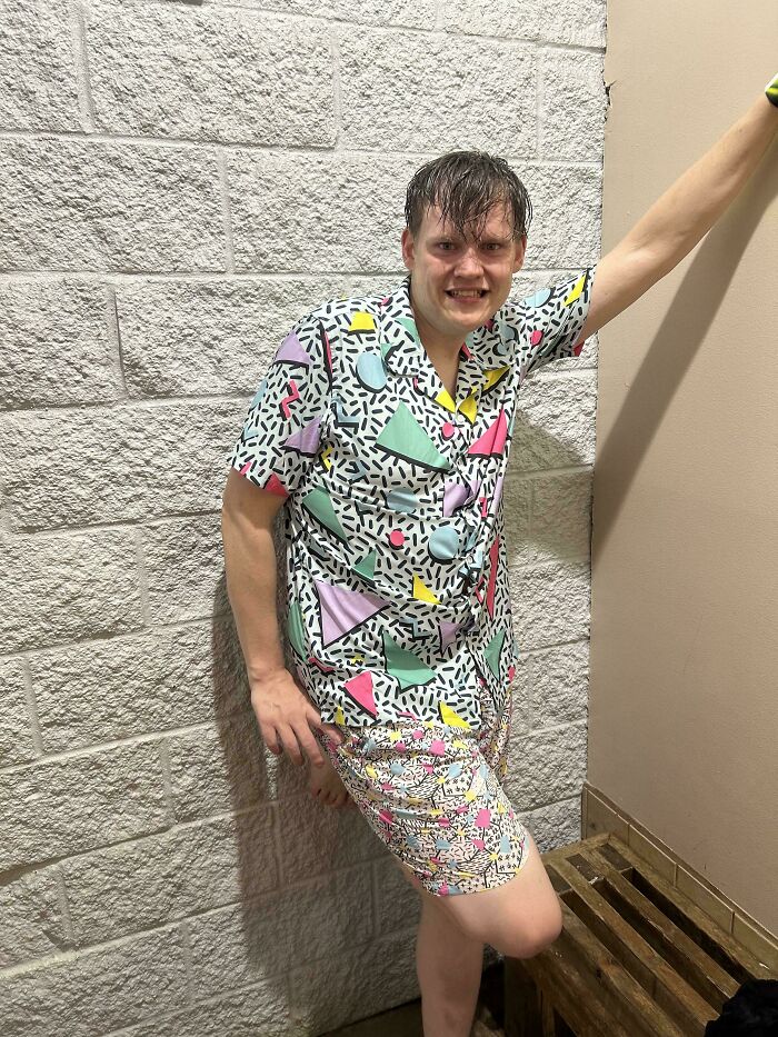 Young man in colorful 90s-inspired clothes smiling and leaning against a brick wall, evoking nostalgic 90s vibes.