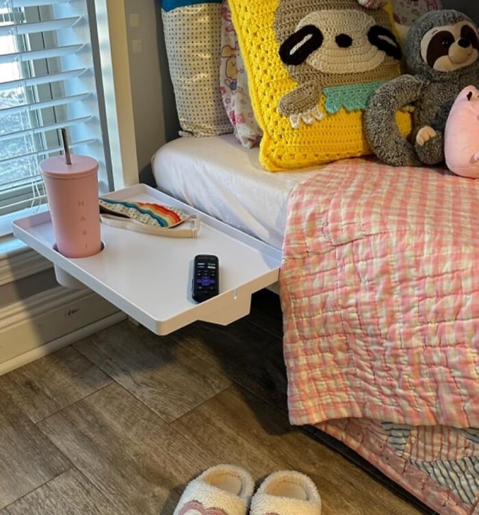 Wall-mounted tray table with a pink cup, mask, and remote beside a bed, a smart dorm find for freshman year survival.