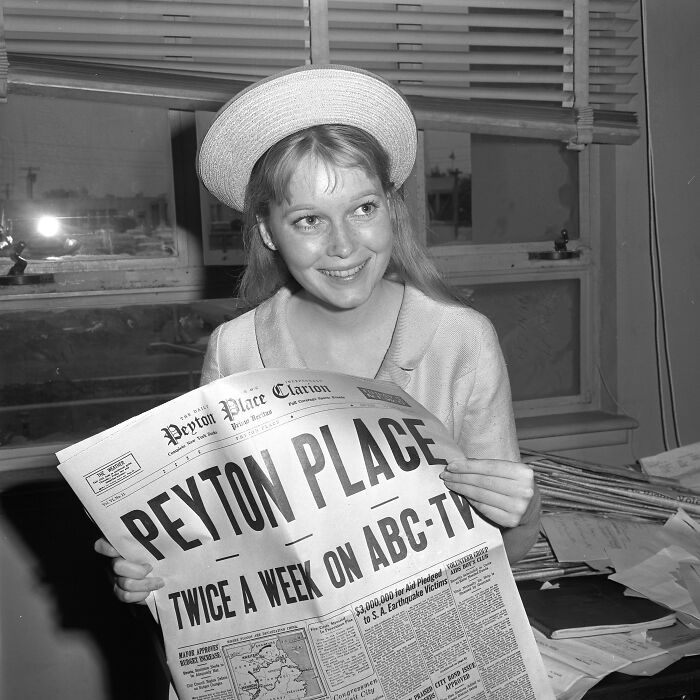 Young LA woman in 1962 smiling indoors, holding a newspaper, capturing moments of women living their best lives.