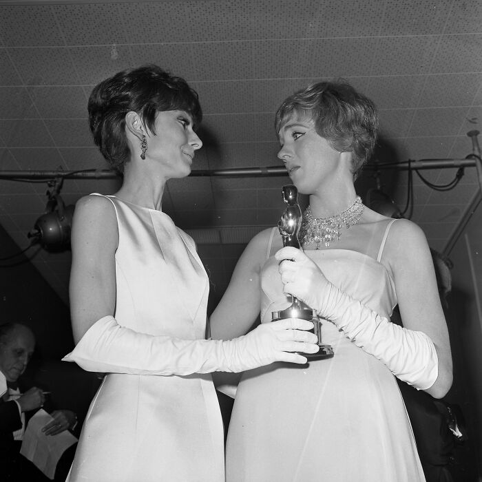 Two LA women in elegant 1960s gowns and gloves, celebrating while holding an award at a glamorous event.