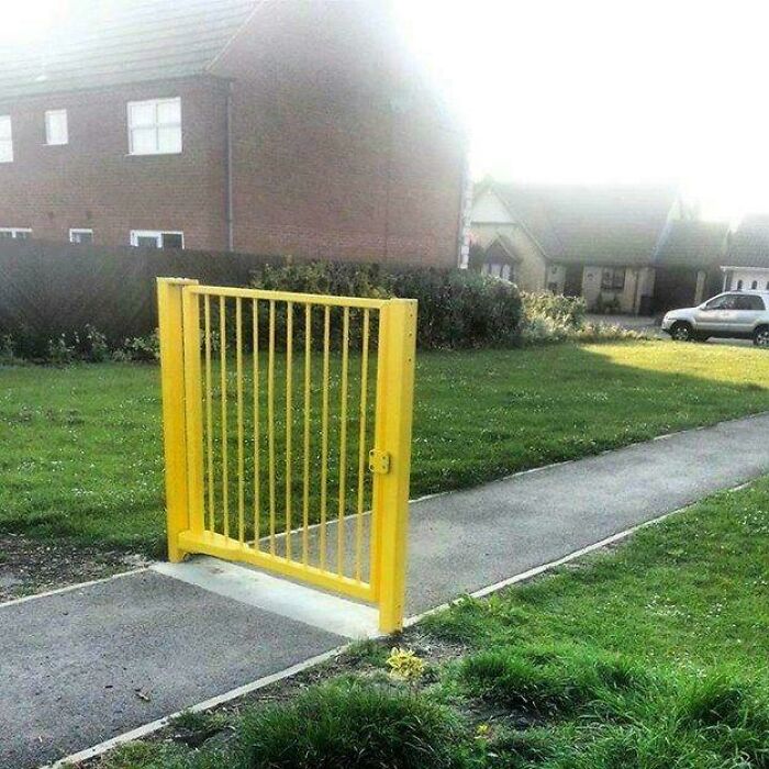Yellow gate standing alone in a British neighborhood, blocking a sidewalk path with houses and a car in the background.