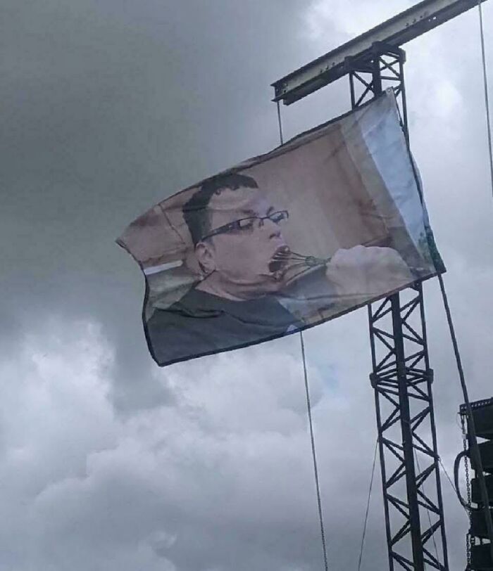 Flag featuring a chaotic British meme of a man eating with a whisk, hanging on a metal structure against a cloudy sky.