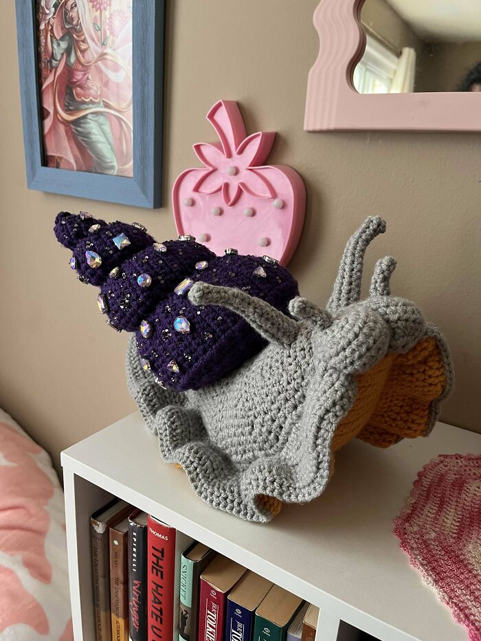 Crochet snail sculpture with detailed shell adorned with beads, displayed on a white shelf in a cozy room setting.
