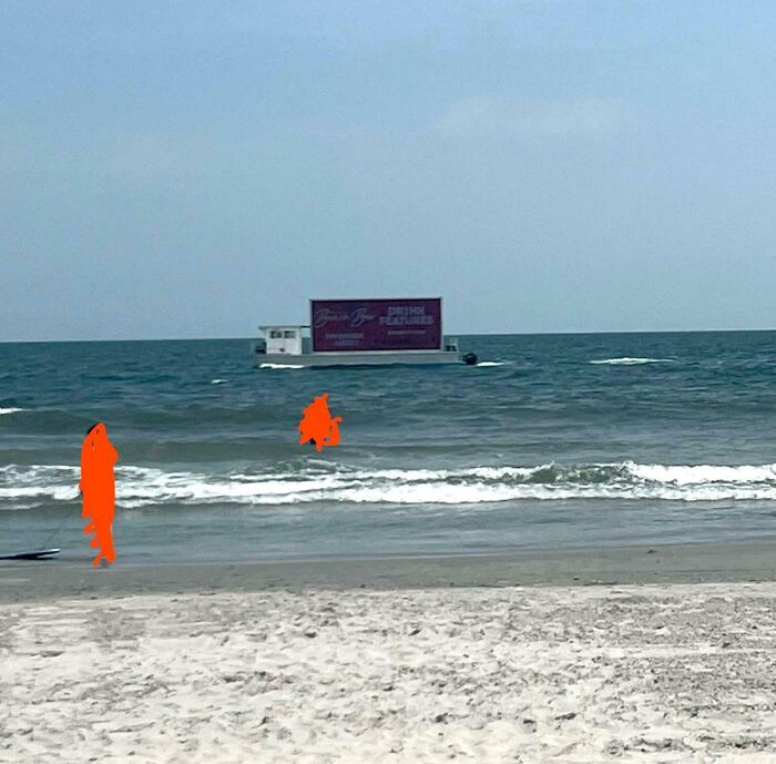 Boat with large advertisement floating near beach waves, illustrating impact of consumerism on natural environments.