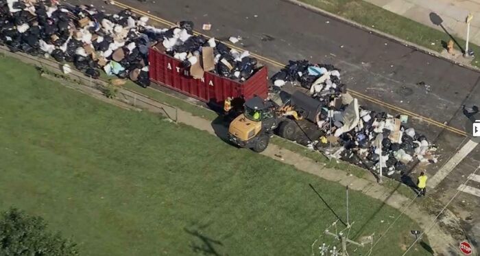 Aerial view of large piles of consumer waste on a street with a loader clearing trash, illustrating consumerism impact.