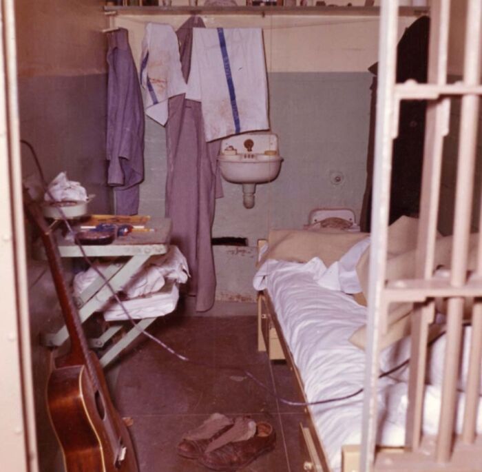 Prison cell interior on Alcatraz showing worn shoes, bed with white sheets, hanging clothes, and a guitar leaning against a table.