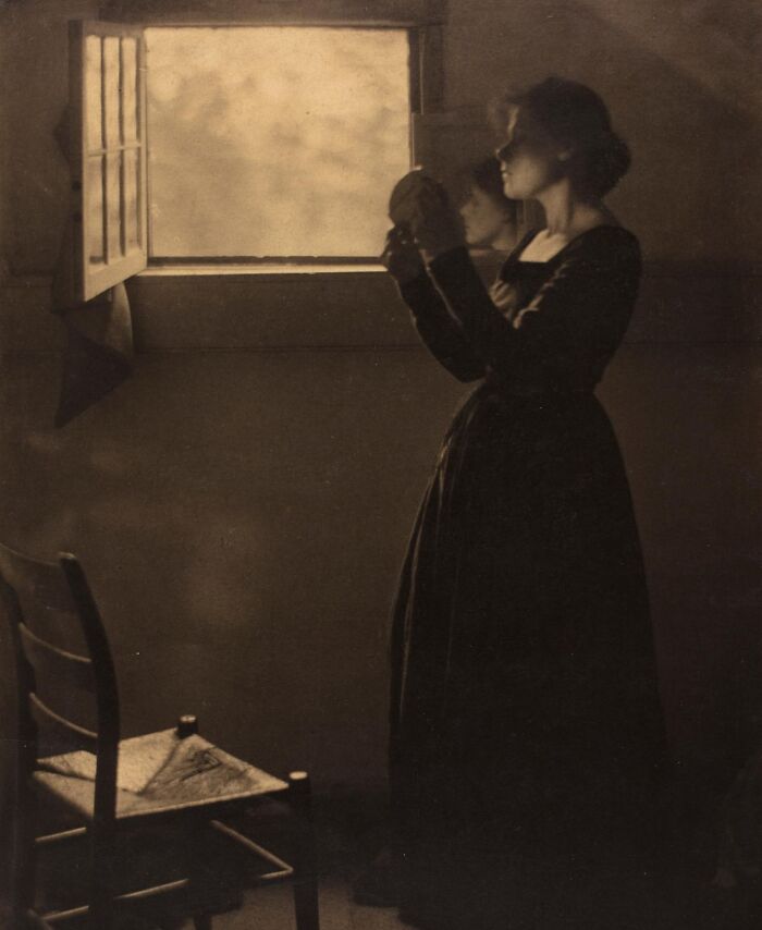 Woman in vintage dress holding an object by an open window in a dimly lit room, reflecting 20th century triumph and tragedy.