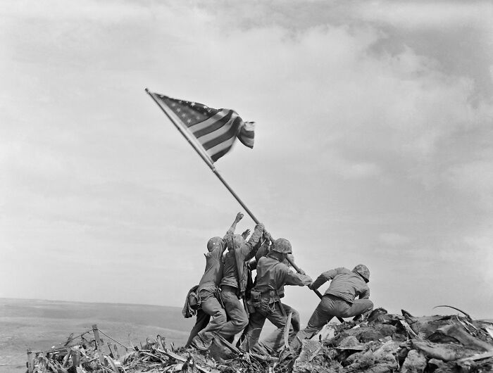 US soldiers raising the American flag on a battlefield hilltop, a symbol of 20th century triumph and tragedy.
