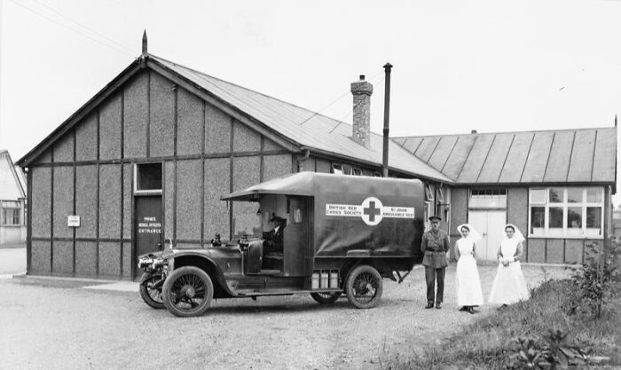 WWI women in nursing uniforms standing next to a British Red Cross ambulance near a wartime medical building.