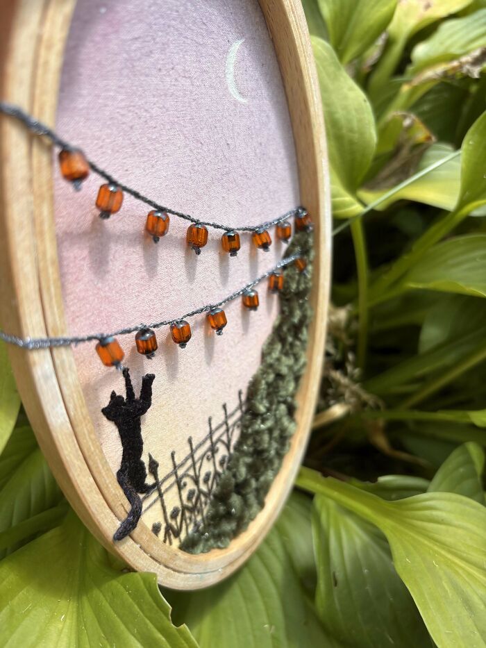 Close-up of beautiful embroidery featuring a black cat, orange beads, and textured green foliage in a wooden hoop.