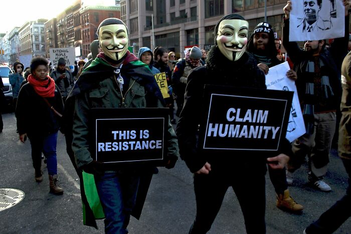 Protesters wearing Guy Fawkes masks holding signs that say this is resistance and claim humanity during a chaotic good demonstration.