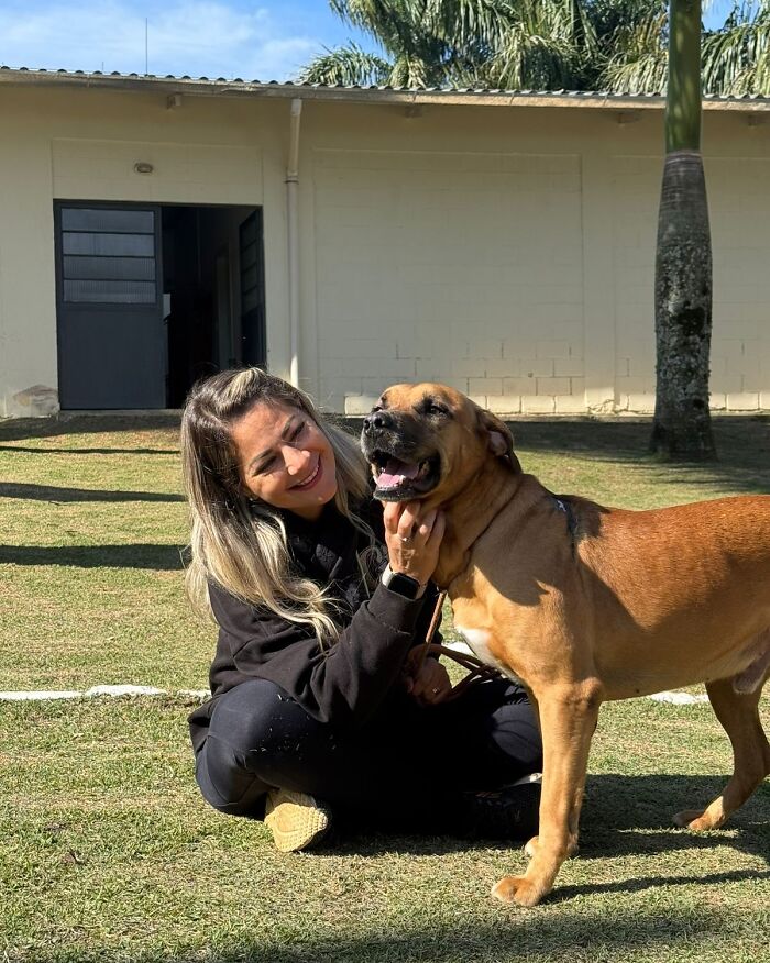 Woman sitting on grass happily petting a shelter dog during joyful weekly walks outside a building.