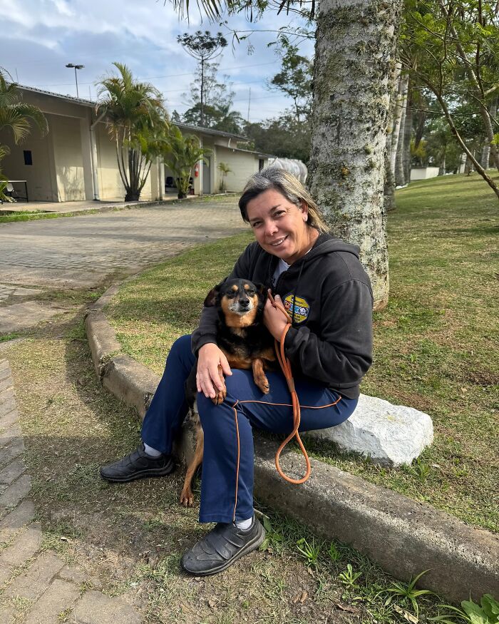 Woman sitting outside holding a happy shelter dog on leash, enjoying a joyful weekly walk in a green park area.