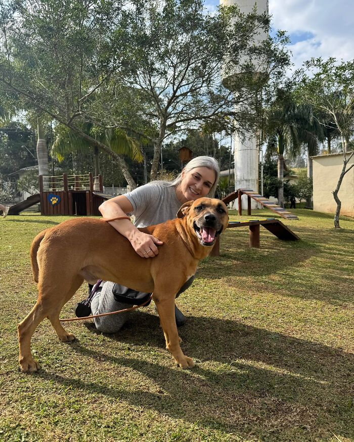 Woman smiling and hugging a happy shelter dog on their weekly walk in a sunny outdoor play area.