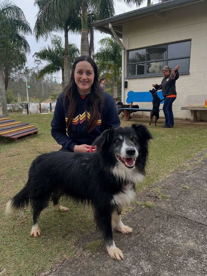 Woman smiling with a happy black and white shelter dog outside during weekly walks on a grassy area with palm trees.