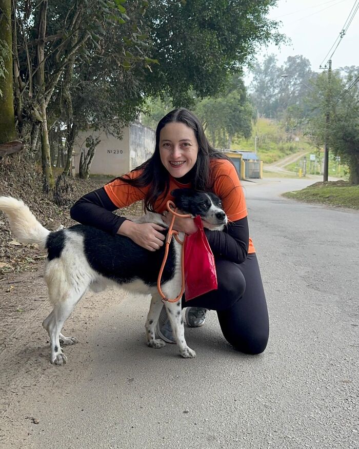 Woman in orange shirt kneeling and hugging a happy black and white shelter dog on a quiet rural road during a walk