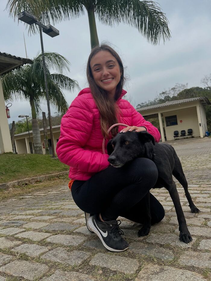 Young woman in a pink jacket kneeling and smiling with a black shelter dog during a joyful weekly walk outdoors.