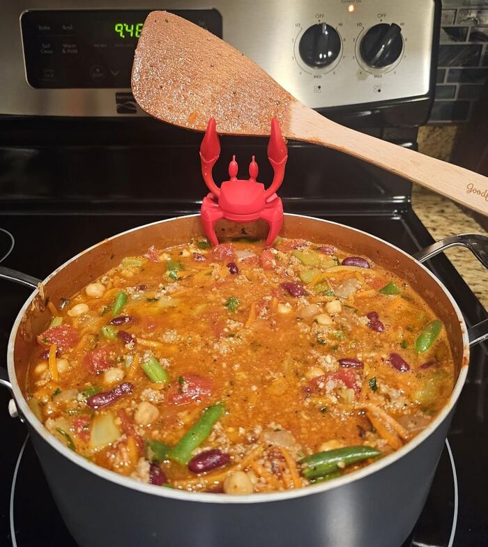 Red crab-shaped kitchen tool holding a wooden spatula above a simmering vegetable and bean stew on the stove.