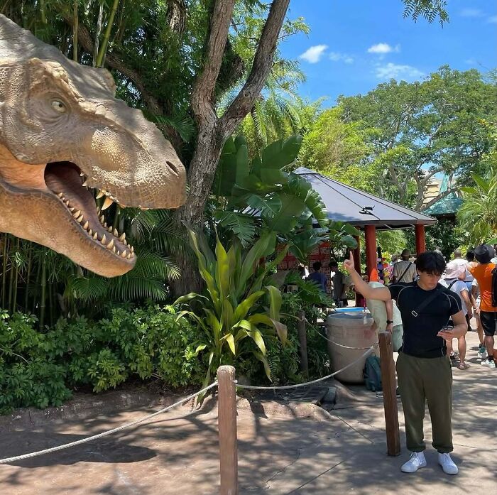 Man posing next to a lifelike dinosaur statue in a tropical park, a funny vacation picture moment.