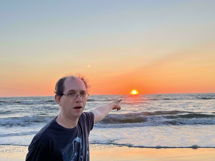 Man with glasses pointing at a sunset over ocean waves during a vacation, a funny and awkward vacation pic moment.