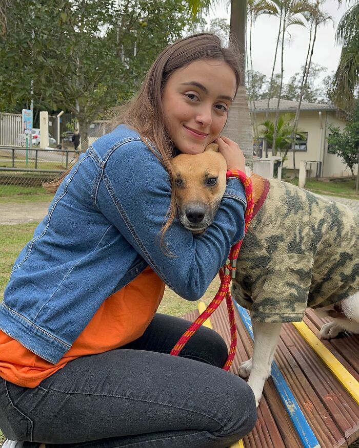 Young woman lovingly hugging a shelter dog wearing a camouflage jacket during a joyful weekly walk outdoors.