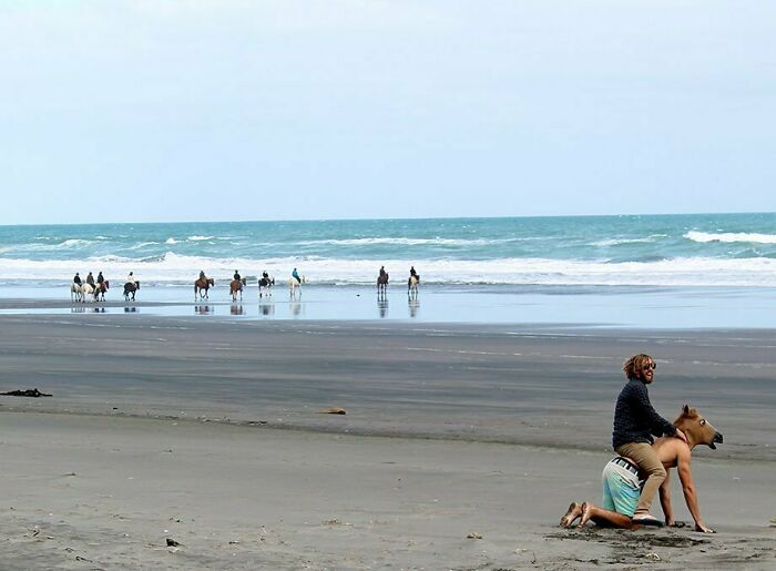 Two men posing for a funny vacation pic on a beach, one wearing a horse mask while kneeling in the sand.