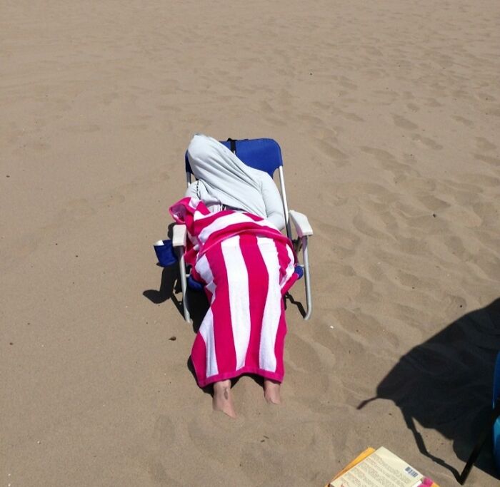 Person wrapped in a towel and hoodie lying in a beach chair on sand, a funny vacation pic from a comedy special series.