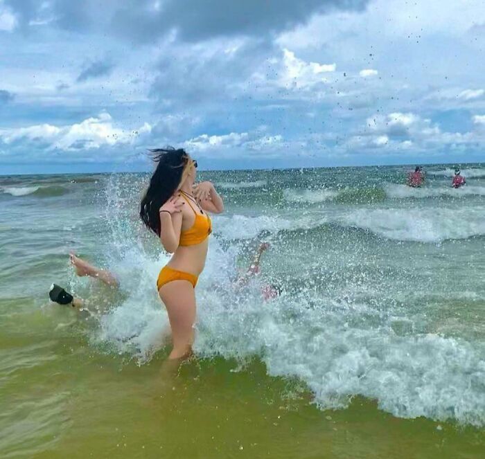 Woman in orange bikini reacting as waves splash a person falling behind her during a funny vacation photo at the beach.