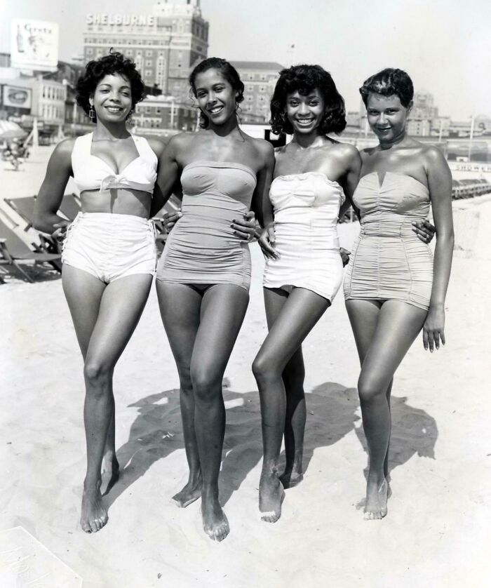 Four women in vintage swimsuits posing happily on a sunny beach, showcasing candid glimpses into the past.
