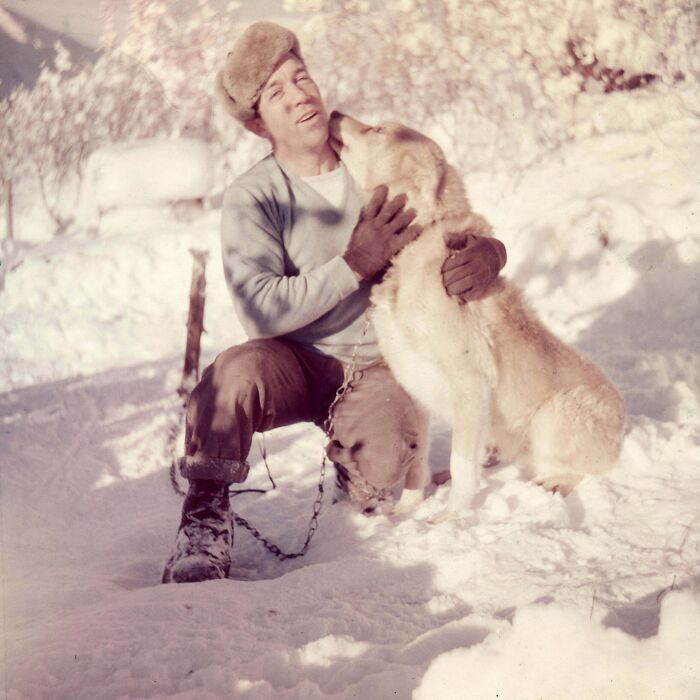 Man wearing fur hat and gloves kneeling in snow, affectionately holding a chained dog in candid glimpses into the past.