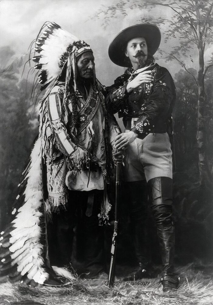 Black and white 19th century photo of a Native American chief and a man in Western attire holding a rifle and posing together.