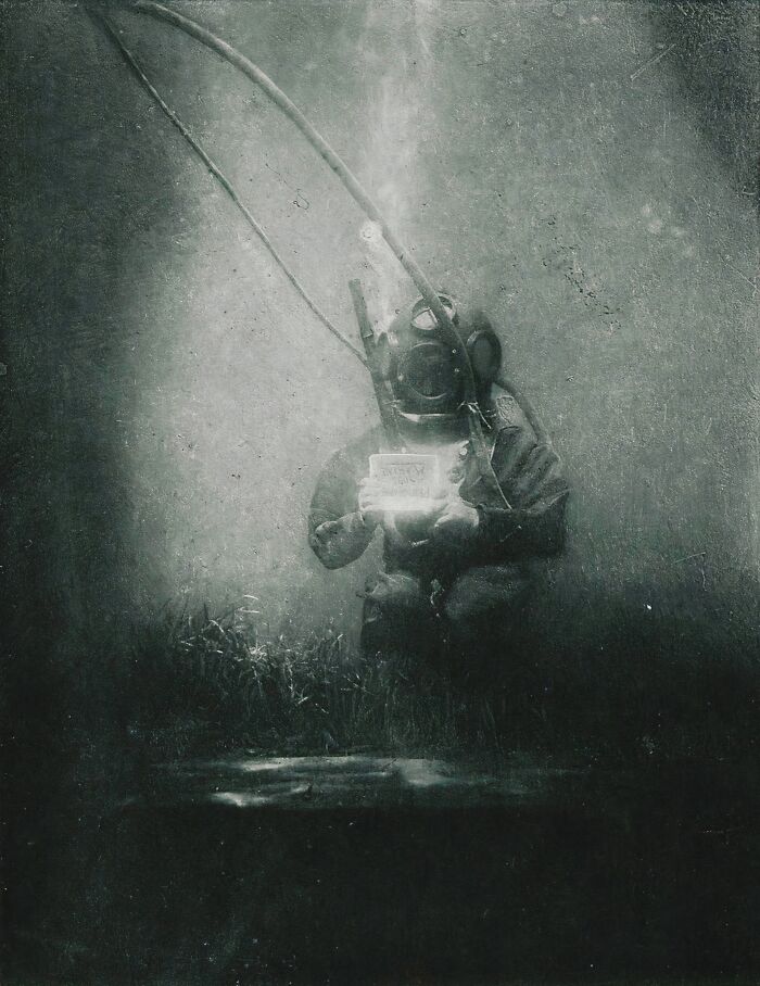 Underwater black and white photo of a 19th century diver in an old-fashioned diving suit holding an object.