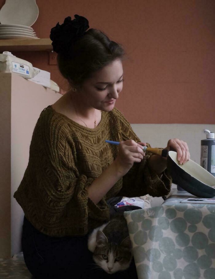 Young woman painting a bowl indoors with a cat resting on her lap, resembling a Renaissance masterpiece scene.