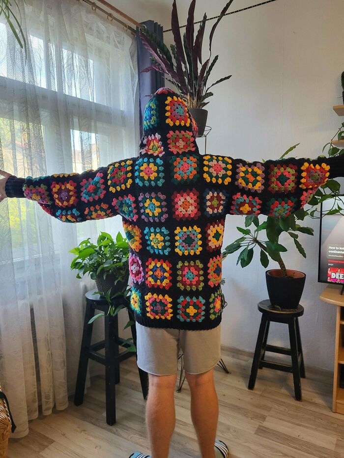 Person wearing a colorful crochet hoodie made of granny squares standing in a bright room with plants and curtains.