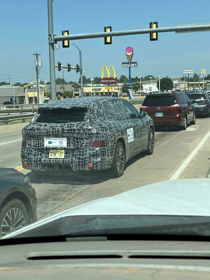 Camouflaged test vehicle on a busy street with recognizable fast food signs in the background, moderately interesting scene.
