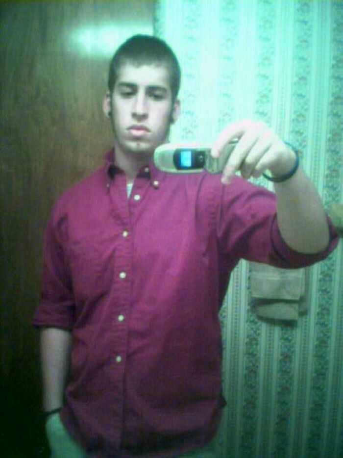 Young man in a red button-up shirt taking a mirror selfie with an old flip phone, a cringe-worthy regrettable past moment.
