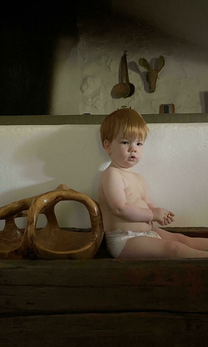 Toddler with red hair sitting on rustic wooden bench in dim light, an accidental renaissance photo resembling classical painting style.