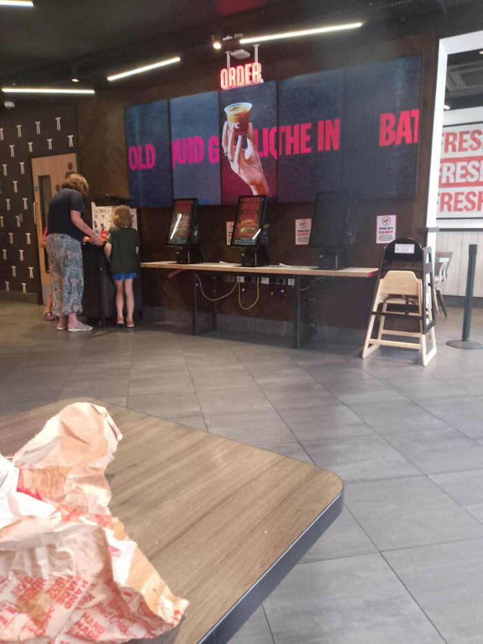Fast food restaurant self-order kiosks with a partially broken digital menu and people standing nearby, illustrating a preventable fail.