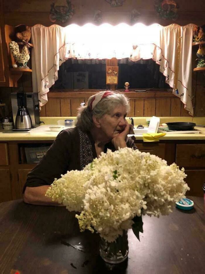 Elderly woman leaning on hand in a rustic kitchen with white flowers, an accidental Renaissance photo moment.