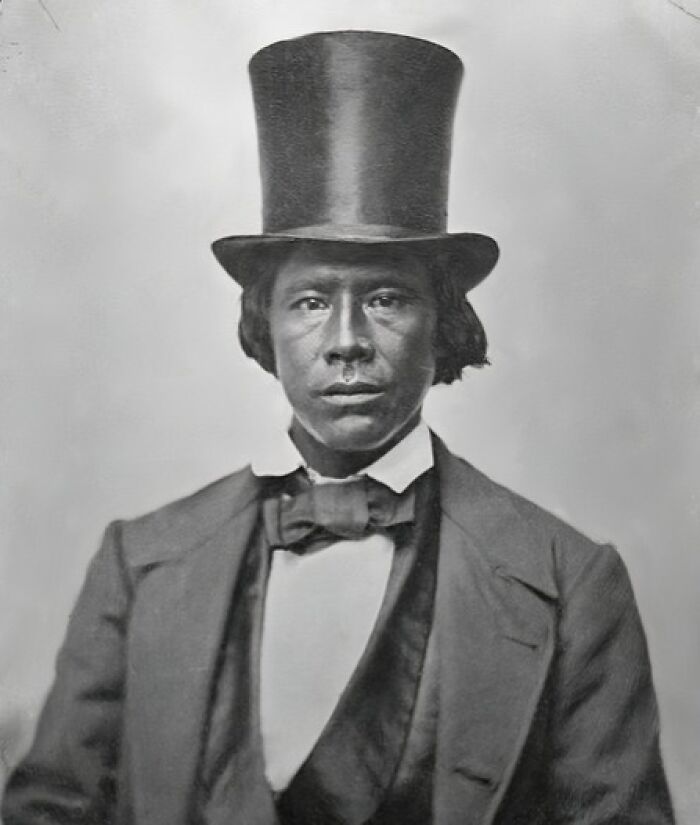 19th century historical photo of a man wearing a tall top hat and formal suit in a vintage portrait.