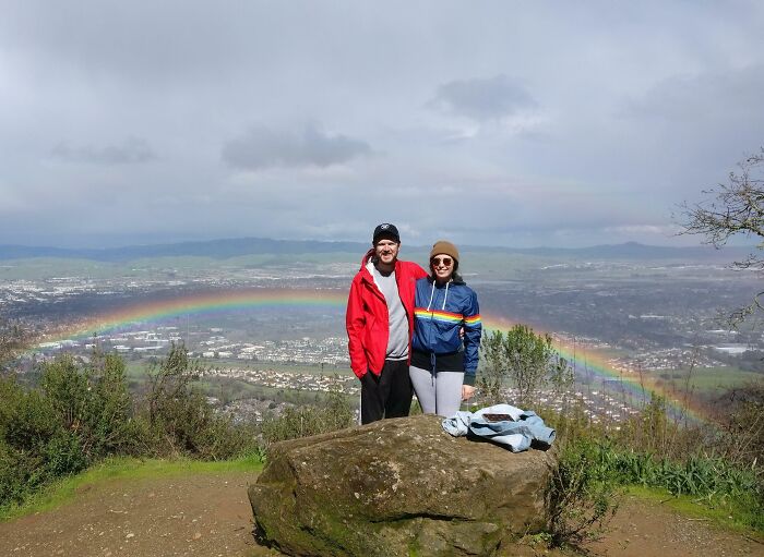 Couple posing on a hill with a rare rainbow coincidence perfectly framing them over a cityscape under cloudy skies