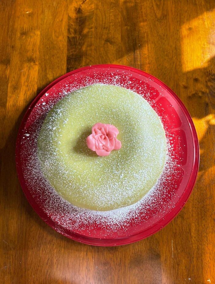 Round baked good dusted with powdered sugar and topped with a pink frosting rose on a red plate on wood table.