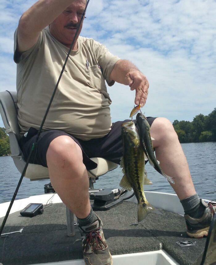 Man sitting on boat holding two fish caught with a single fishing lure, a rare coincidence in fishing photos.