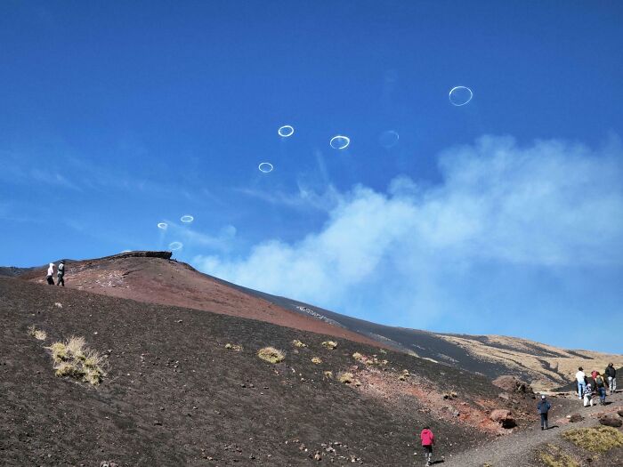 Rare coincidences of smoke rings appearing in a clear blue sky above a volcanic hillside with hikers nearby.