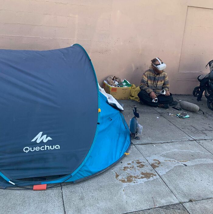 Man wearing virtual reality headset sitting on sidewalk near a tent and belongings in a point of no return photo.