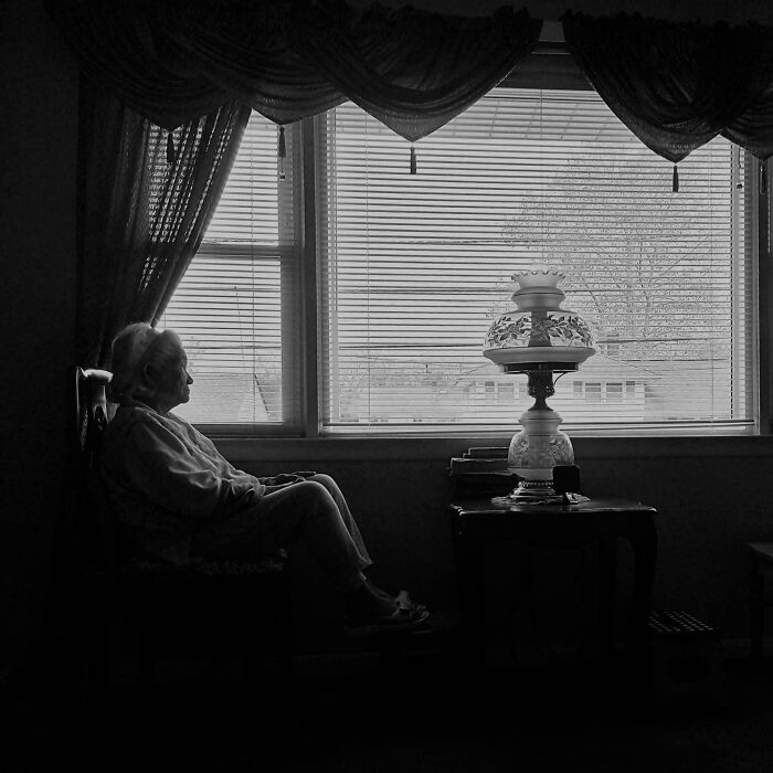 Elderly person sitting by window with vintage lamp, creating a scene resembling a Renaissance masterpiece photograph.