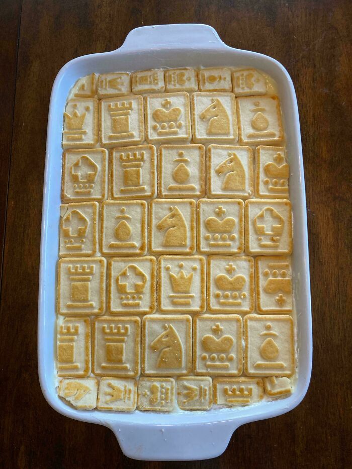 Baked goods arranged with chess piece designs on biscuits in a white baking dish on a wooden table.