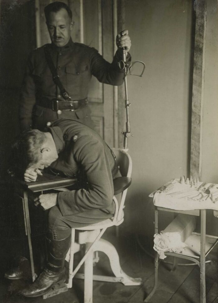 WWI soldier seated with head down while another soldier stands holding a bayonet, revealing daily life beyond trenches.