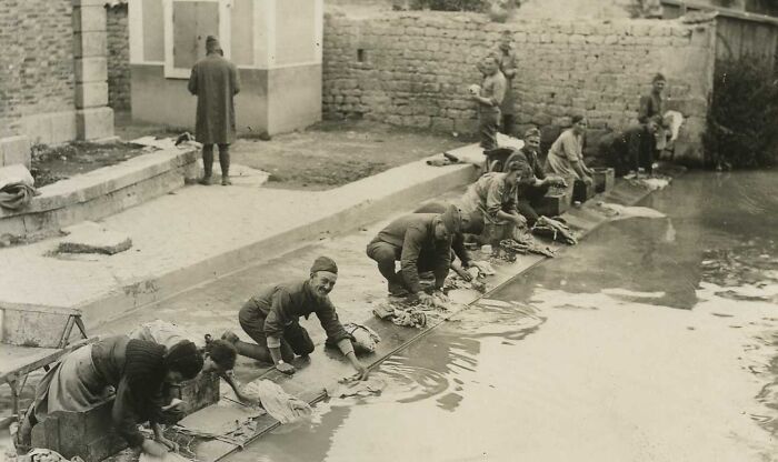 Soldiers washing clothes by a riverbank, showing daily life beyond the WWI trenches in a village setting.